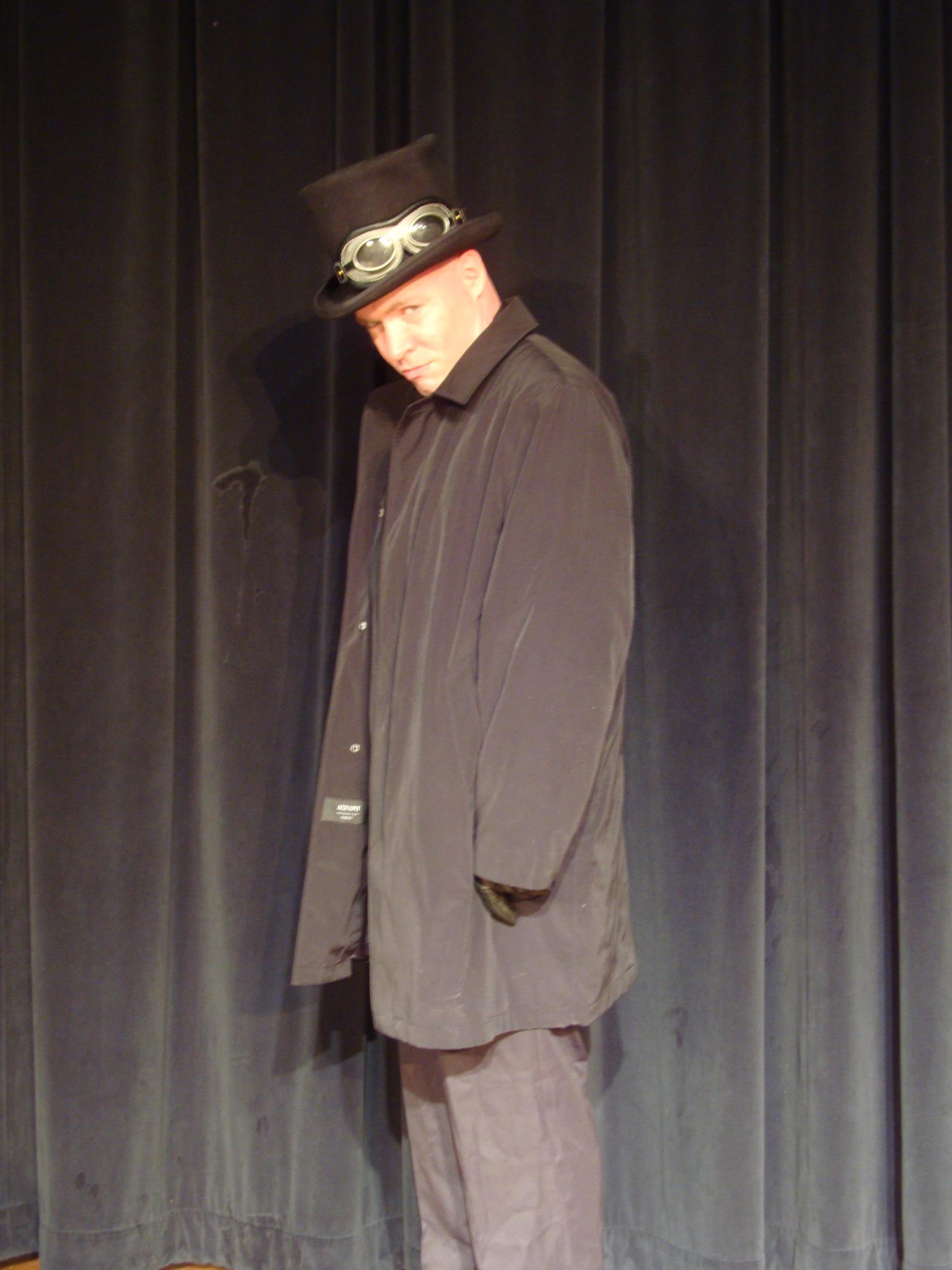 White man in black trenchcoat and black tophat standing against a black curtain